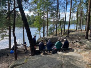 Viisi henkilöä istumassa rantamaisemassa pöydän äärellä ulkoiluvaatetuksessa. Kasvoja ei näy.