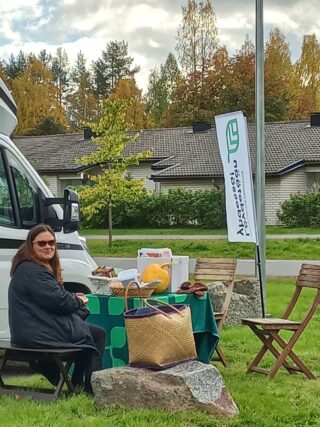Noin 5o-vuotias ruskeatukkainen nainen istuu oöydän ääressä, jossa on esillä syksyn juures- ja kasvissatoa. Takana pakettiauto ja lippu, jossa lukee Löydetään yhdessä.