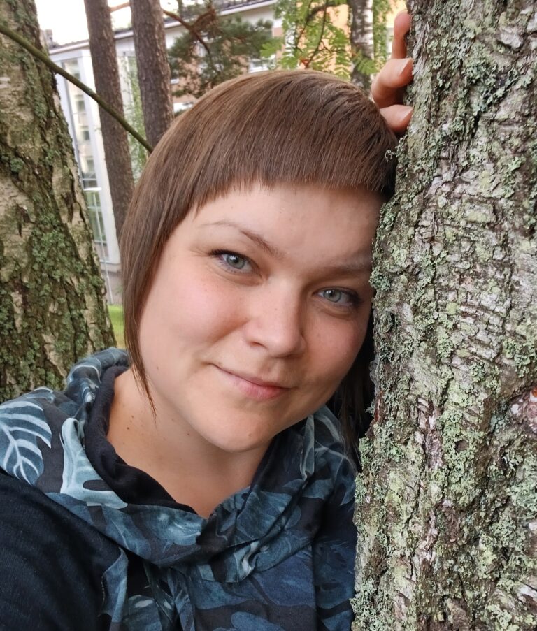 Ruskea tukkainen naishenkilö katsoo kameraan ja nojaa puun runkoon.