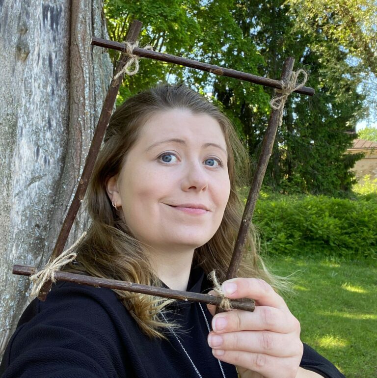 Nainen pitelee kädessään oksista tehtyä kehystä jonka keskellä näkyvät hänen kasvonsa. Naisella on tummat pitkät hiukset jonka latvoissa on vaaleaa ja hän hymyilee.